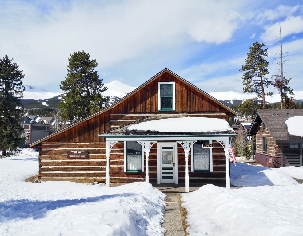Snowy wooden cabin with a white porch in a mountain town under a blue sky, evergreen trees in the background.