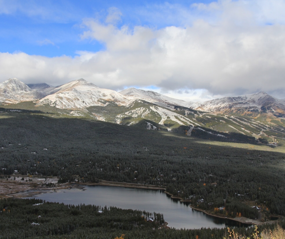 Snow-covered mountains under a blue, partly cloudy sky with a pine forest and a calm lake in the foreground (aerial view).