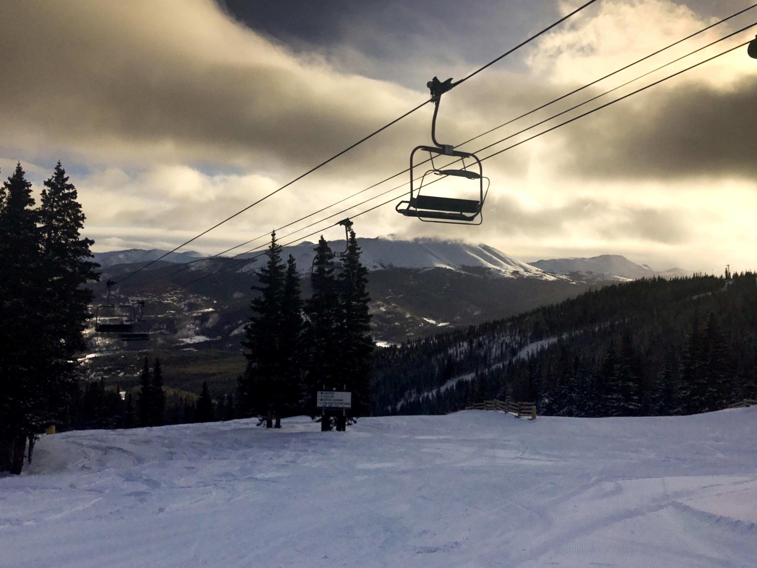 An empty ski lift chair suspended on cables above a snowy mountain landscape with pine trees and cloudy sky.