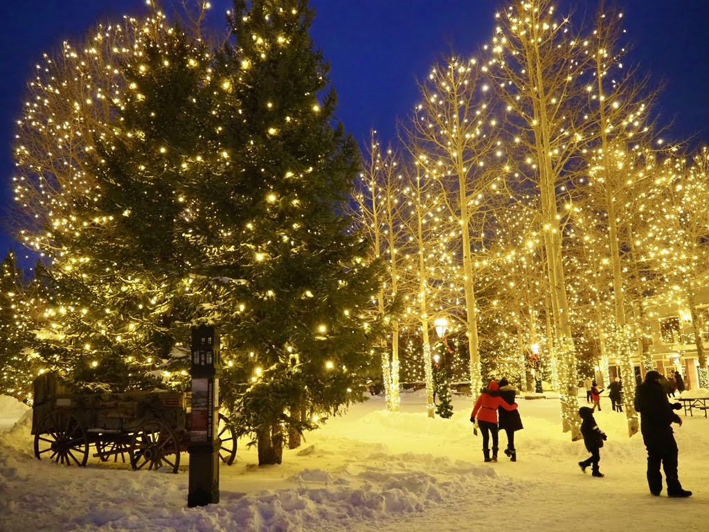 Snowy park at night with trees wrapped in warm holiday lights; people stroll and a couple walks arm-in-arm among glow.