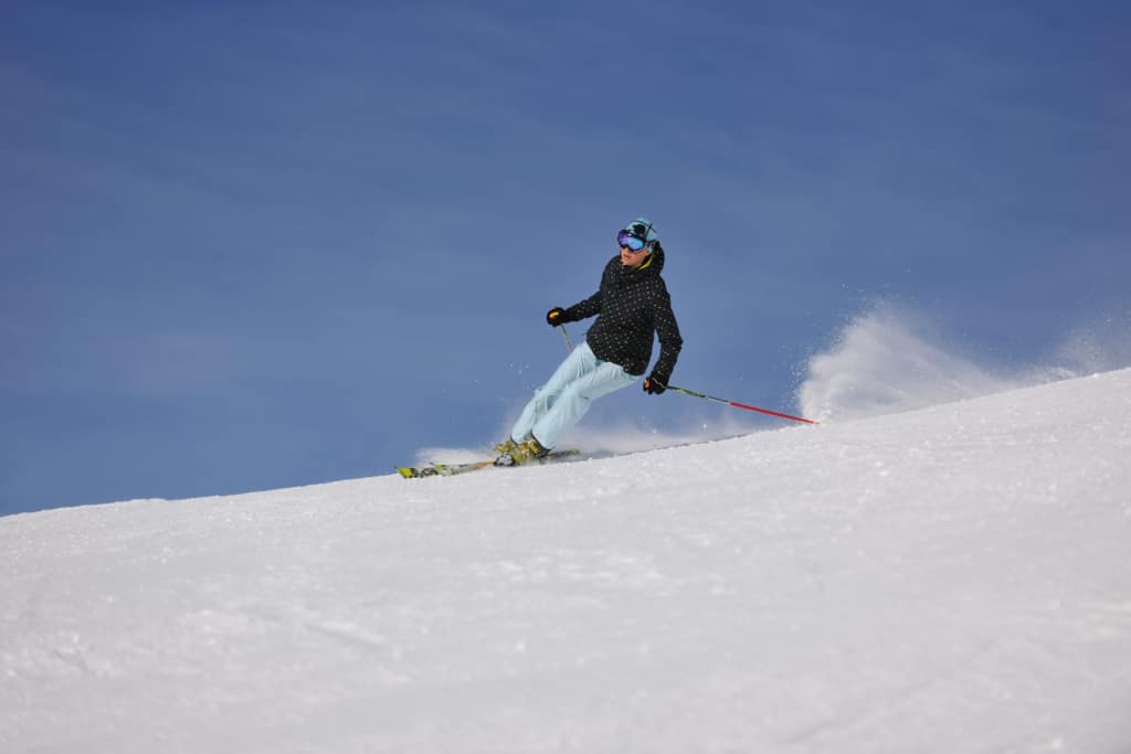 Skier in a dotted jacket and light blue pants makes a turn on a snowy slope under a clear blue sky, wearing goggles and a checkered beanie.