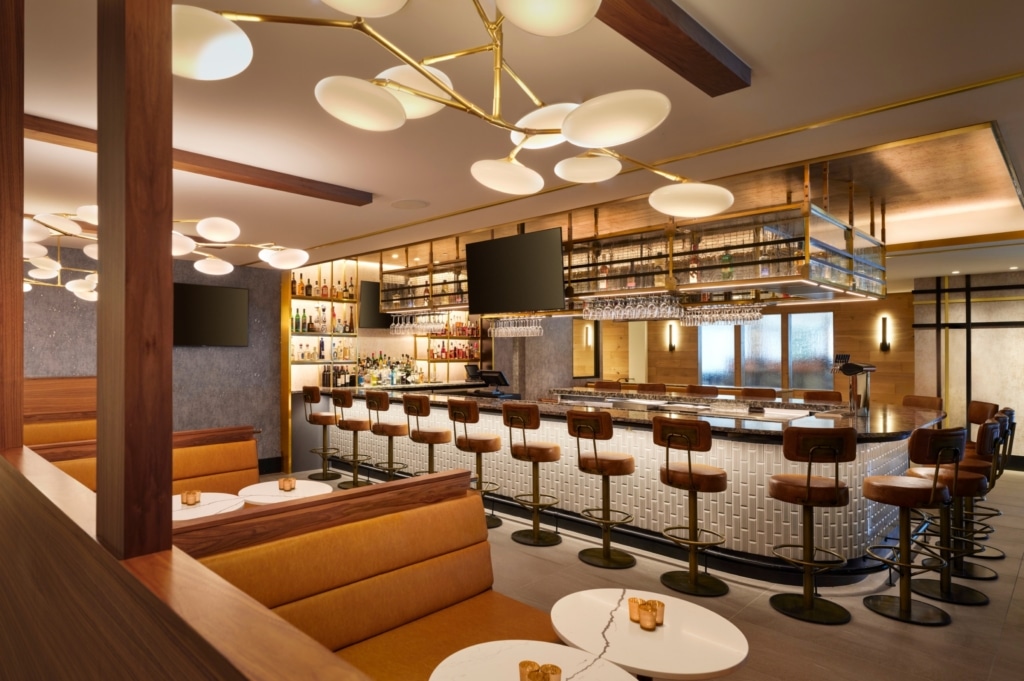Modern bar interior with brass accents, round white pendant lights, and brown leather bar stools along a marble counter.