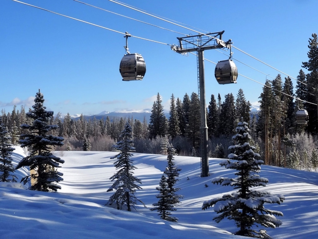 Gondola lift cabins travelling along cables over a snow-covered forest under a clear blue sky at a ski resort.