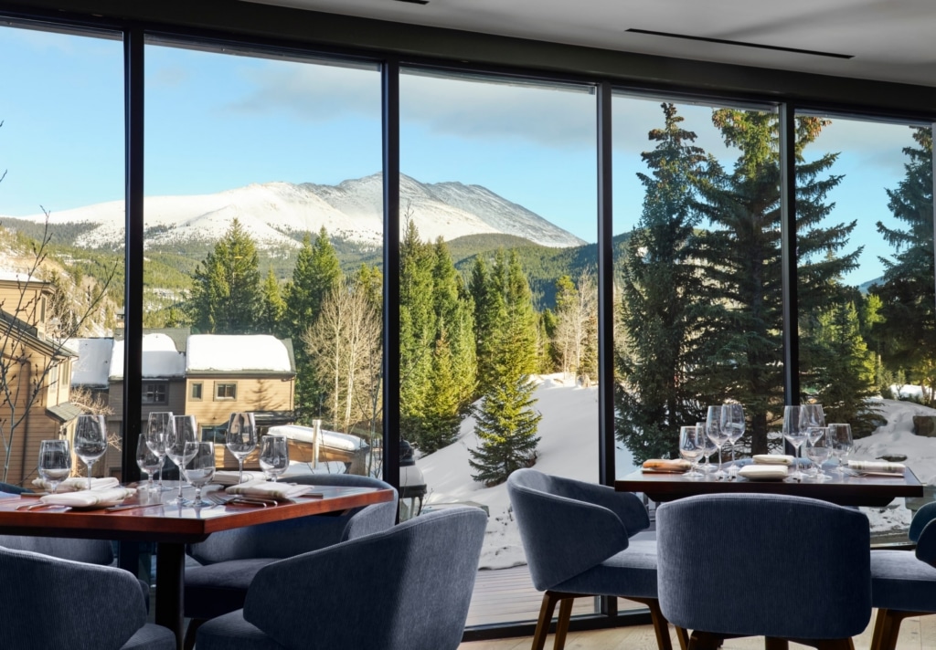 Restaurant dining area with floor-to-ceiling windows and a snowy mountain view outside, tables set with wine glasses and plates.