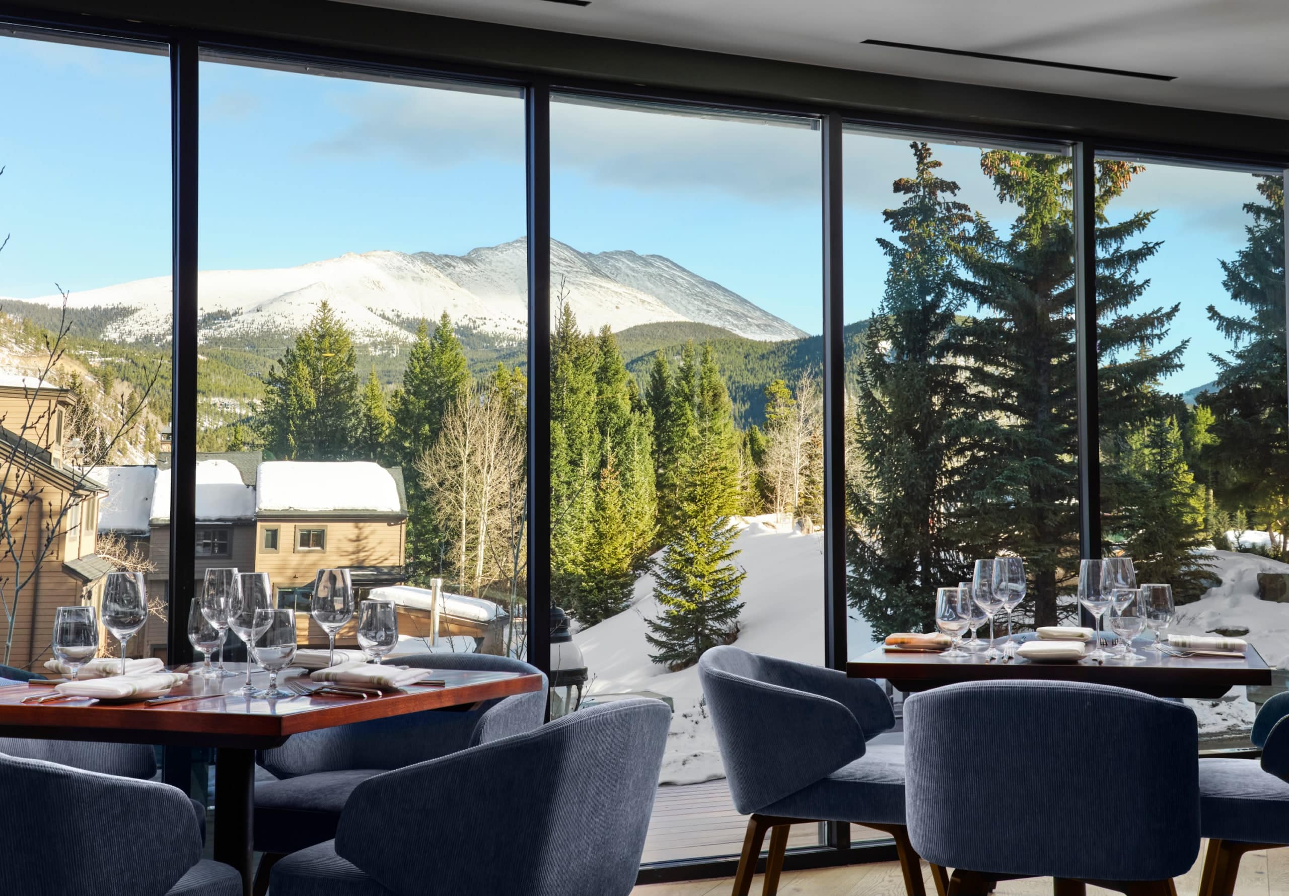 Restaurant dining area with floor-to-ceiling windows and a snowy mountain view outside, tables set with wine glasses and plates.