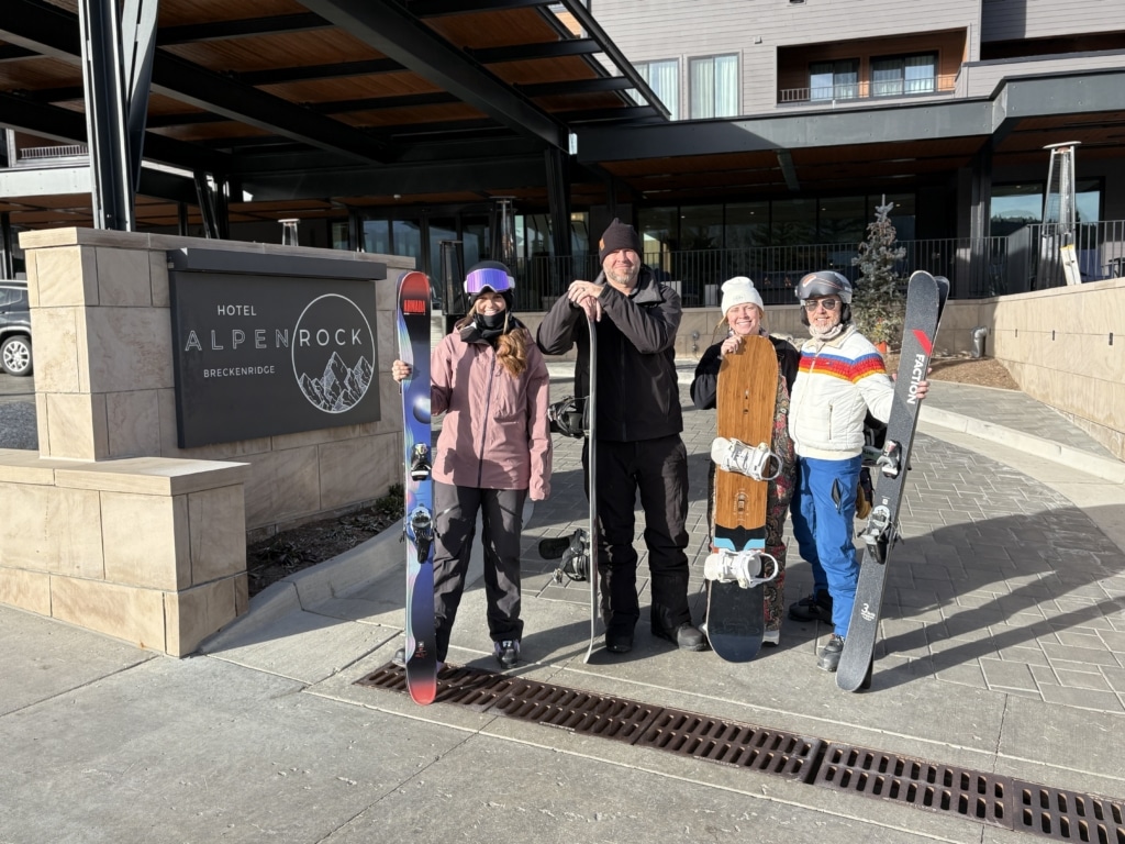 Group of four skiers/snowboarders posing outside a hotel entrance with their gear and boards, Alpine Rock Breckenridge sign visible.