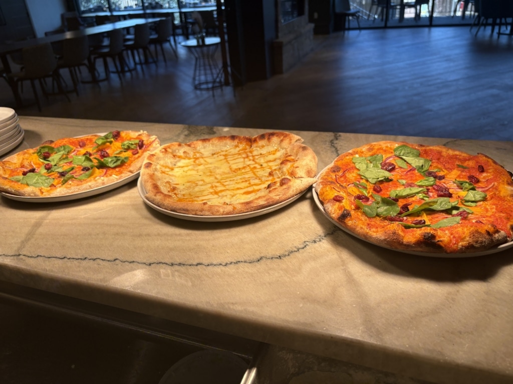 Three pizzas on a marble counter in a dim restaurant, left and right topped with greens and red toppings, center cheese pizza; shelves and seating blurred in background.