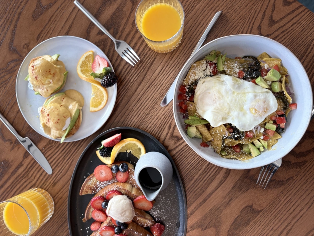 Brunch on a wooden table: waffles with berries, orange juice, and pistachio? no—berries and syrup; eggs Benedict with avocado slices on two plates; orange wedges nearby.