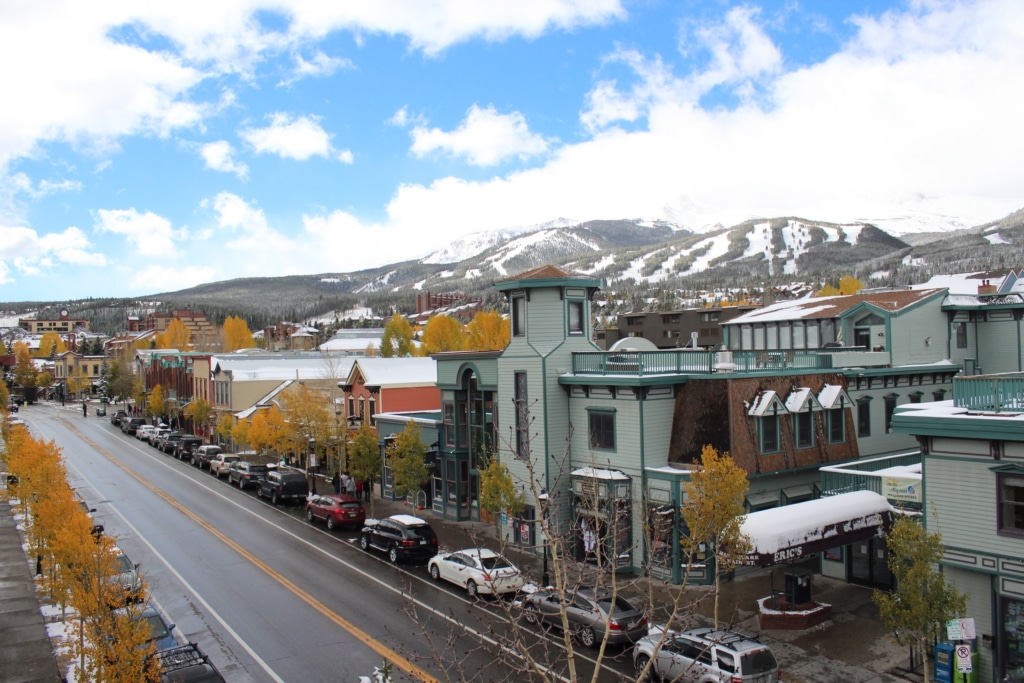 A downtown street with colorful buildings, parked cars, and orange trees, with snow‑capped mountains in the background under a blue sky with clouds.