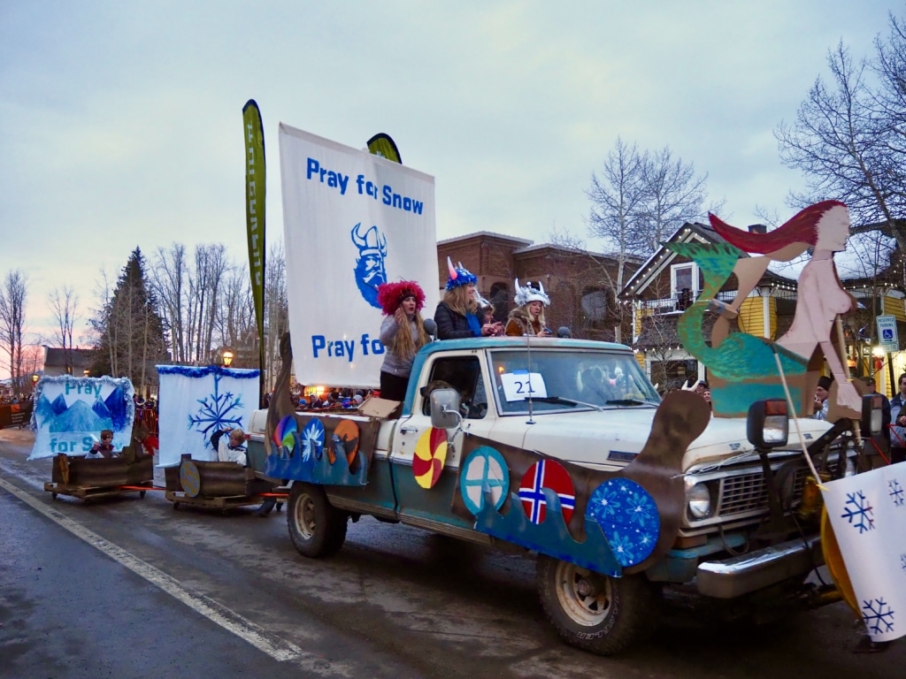 Parade float on a truck with a large banner that says 'Pray for Snow' and people in winter costumes, surrounded by blue snowflake decorations and mountain shapes.