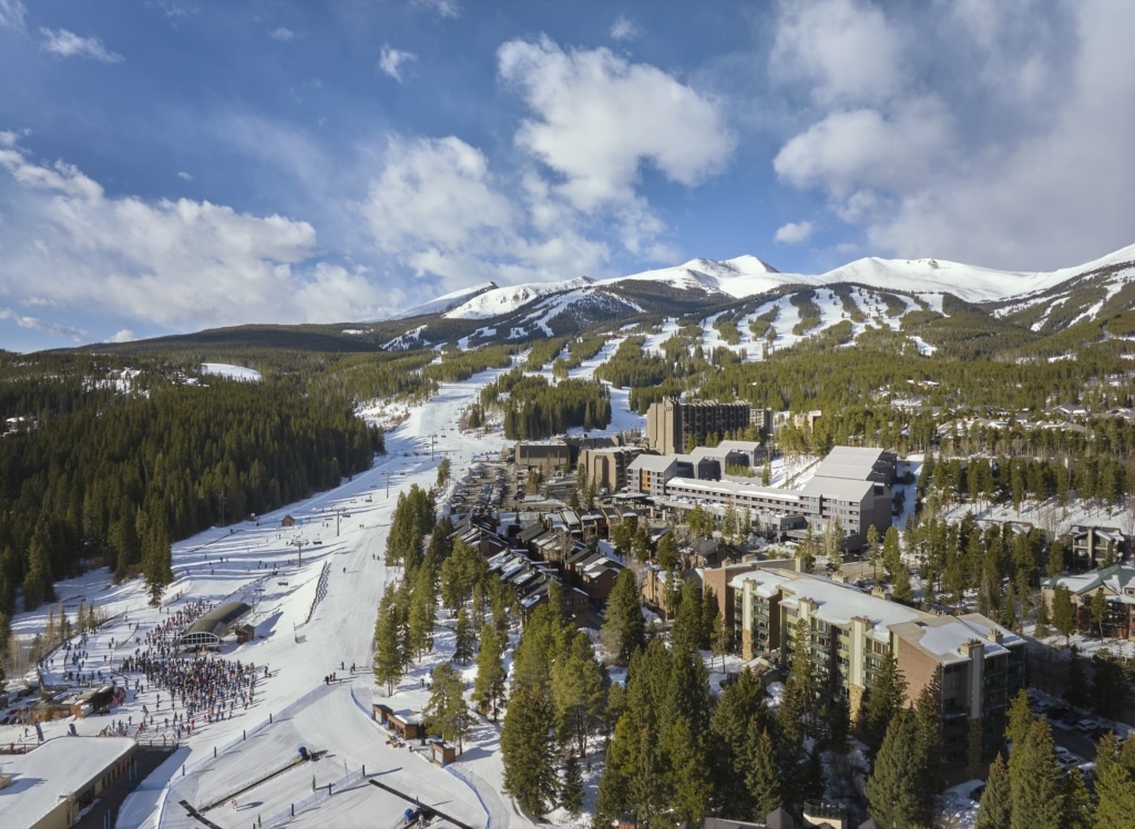 Snow-covered mountain ski resort with groomed slopes, chairlifts, and a cluster of buildings at the base among evergreen trees under a blue sky.