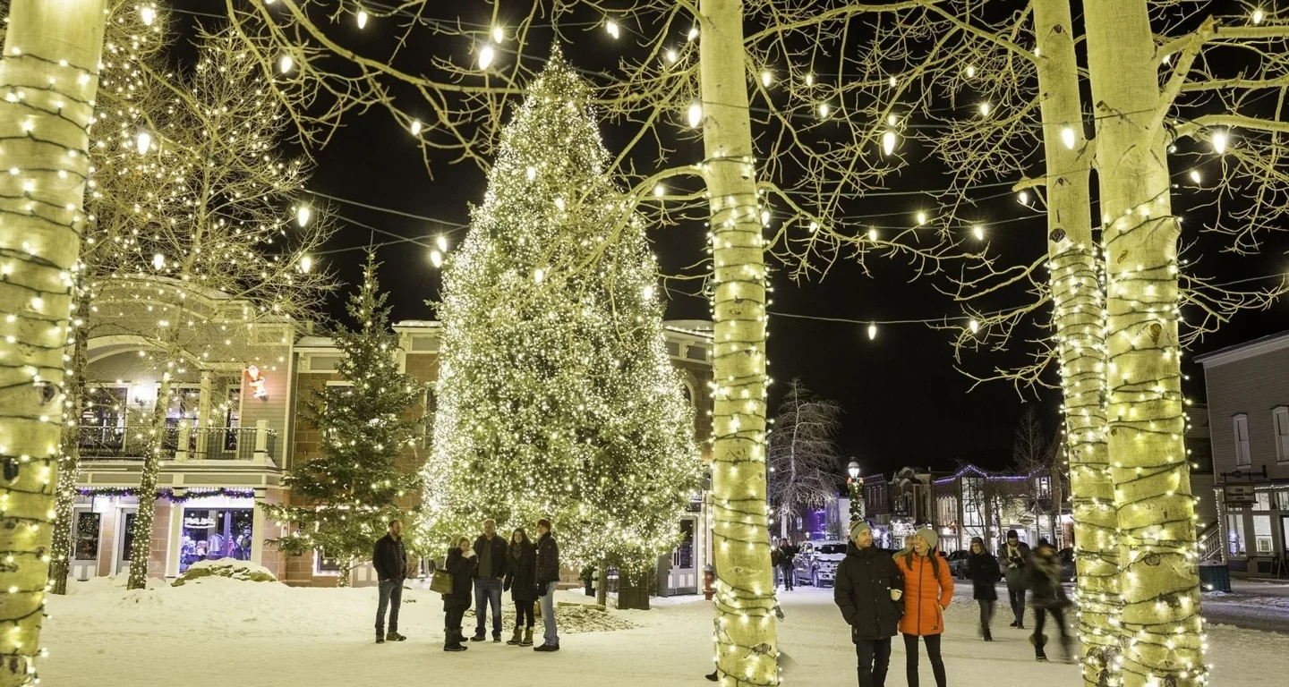 Nighttime street scene with a large illuminated Christmas tree and string lights on bare trees, snow on the ground, and people walking along a festive town plaza.