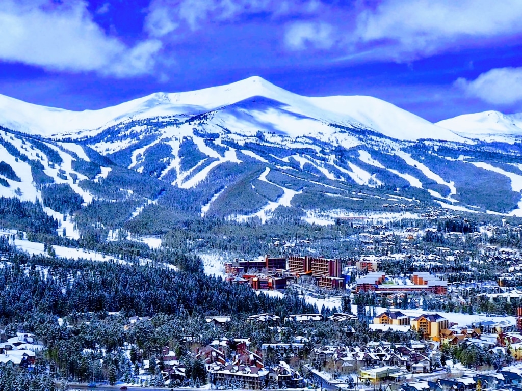 Snow-covered mountain range with a ski resort village at the base, surrounded by evergreen trees under a blue sky.