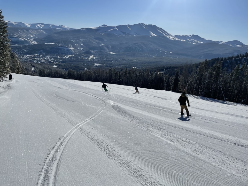 Snowy mountains with skiers on a gentle slope and a clear blue sky; conifer trees line the hillside in the distance.