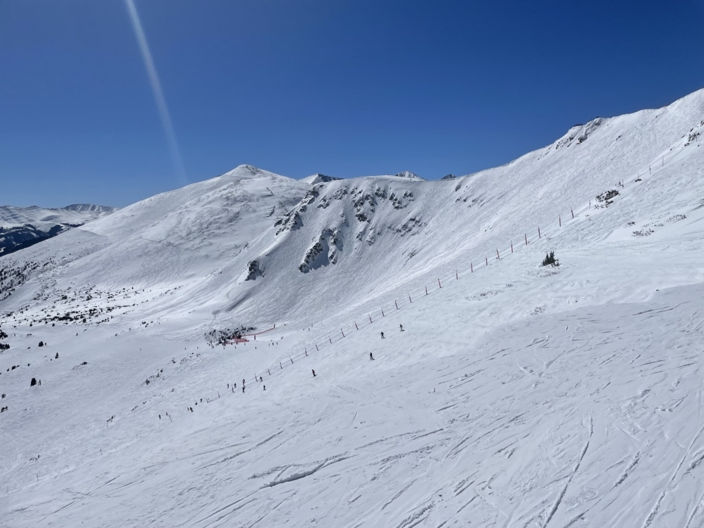 Snowy mountain slope with skiers descending, red safety poles along the hill, under a clear blue sky