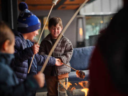 Children in winter jackets roasting marshmallows on sticks over a small flame by a fire pit.